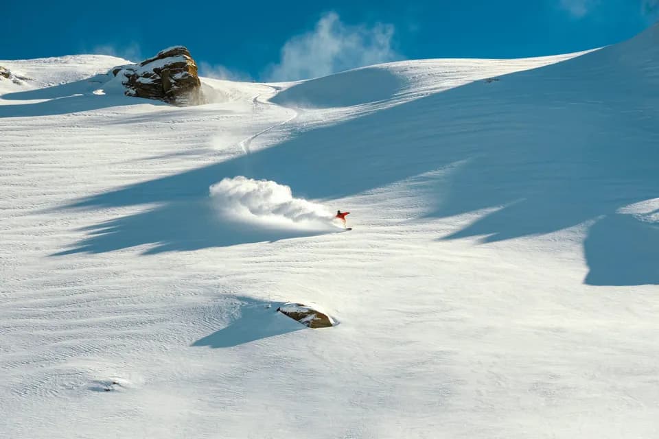 Rider in giacca rossa scende in neve fresca su un ampio pendio fuoripista, sollevando una nuvola di neve