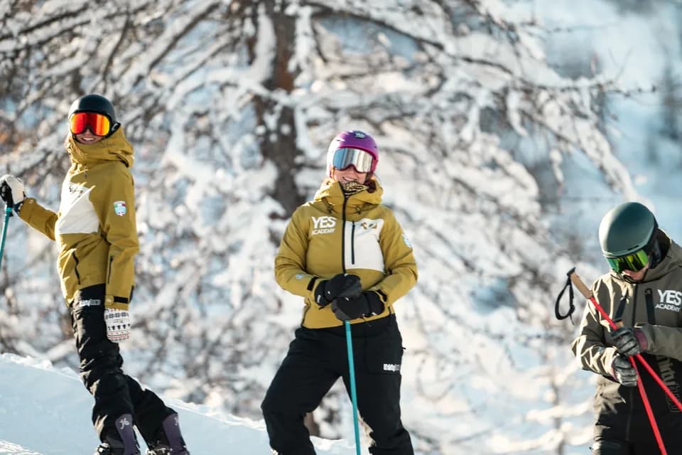 Tre maestri con le giacche YES Academy sorridono in pista davanti a un albero carico di neve