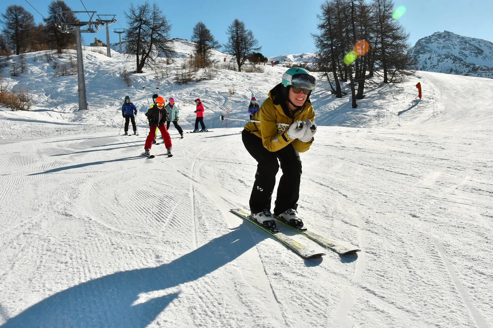 Maestro di sci in posizione a uovo davanti a un gruppo di bambini in fila sulla pista, con seggiovia e larici