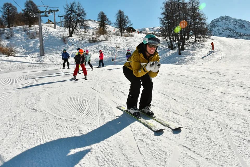 Maestro di sci in posizione a uovo davanti a un gruppo di bambini in fila sulla pista, con seggiovia e larici