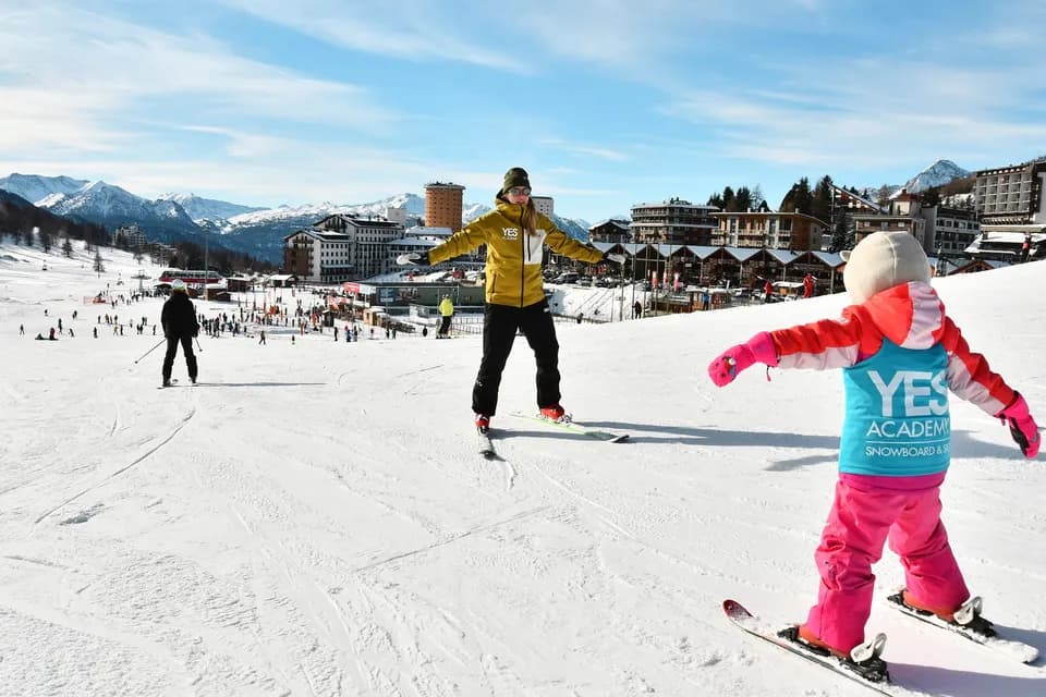 Maestro di sci a braccia aperte guida un bambino piccolo sugli sci con la pettorina YES Academy; sullo sfondo Sestriere