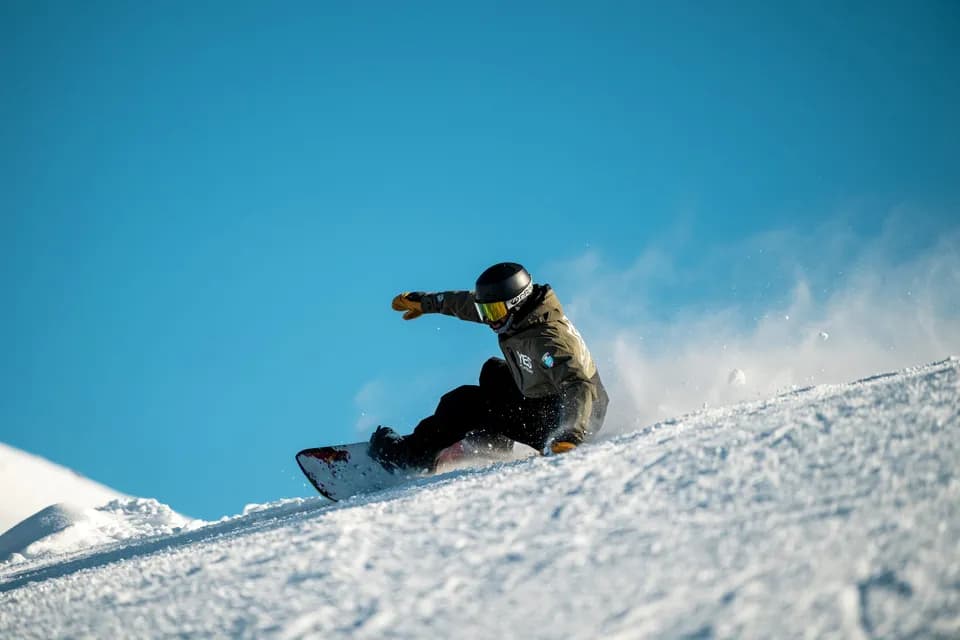 Snowboarder con la giacca YES Academy in curva sulla pista, braccio teso e spruzzo di neve contro il cielo blu