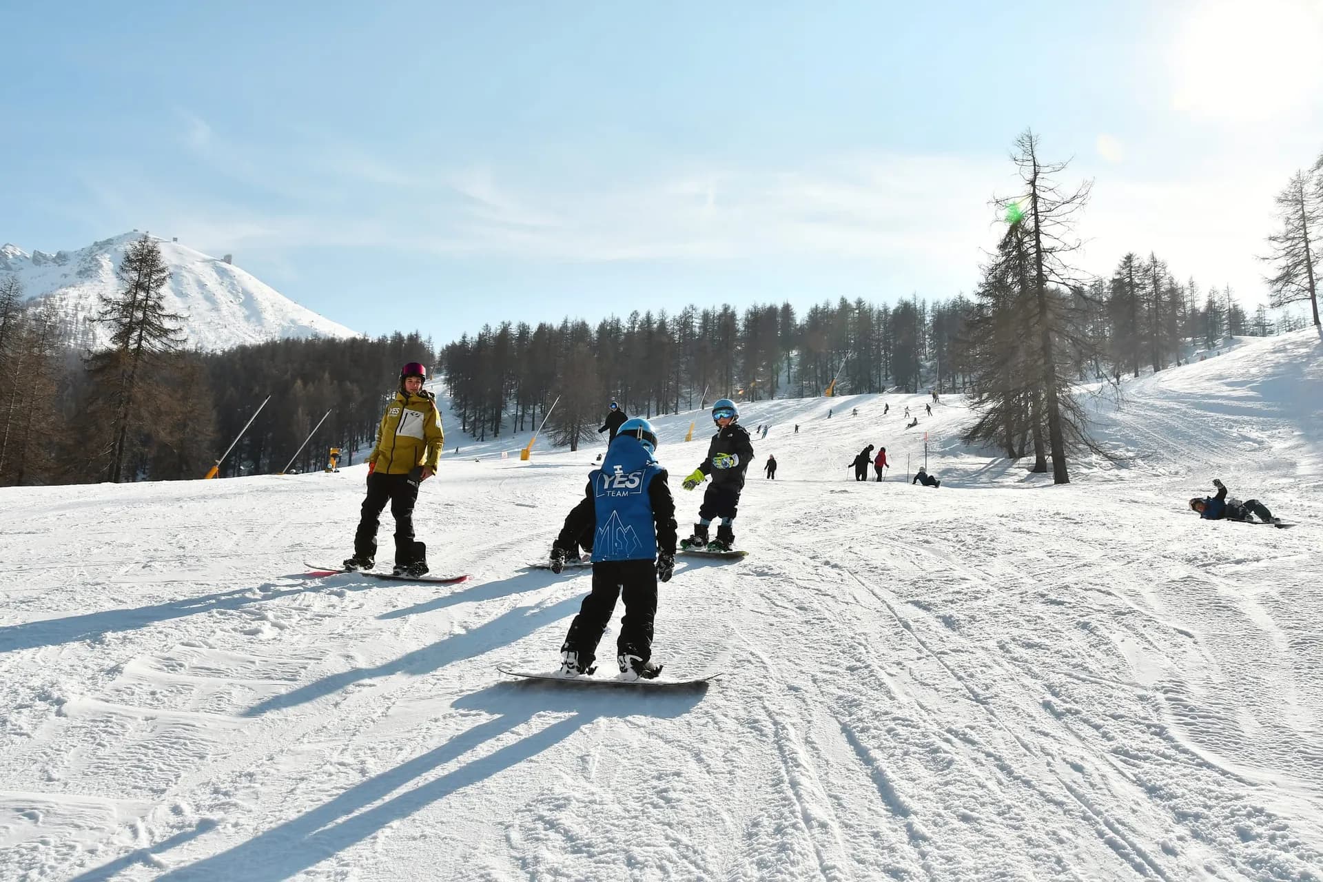 Maestro in giacca gialla guida tre bambini sullo snowboard lungo una pista soleggiata tra i larici