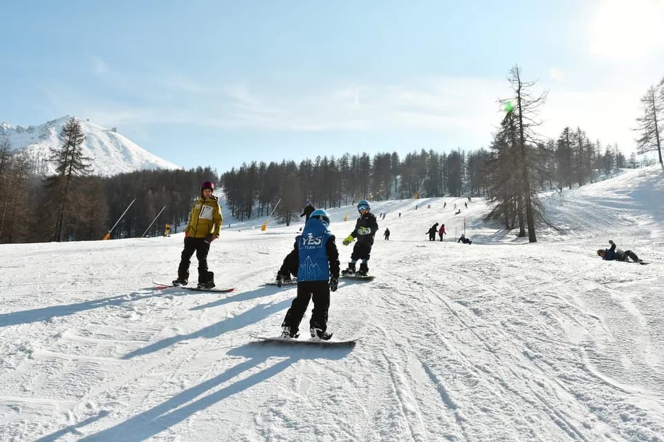 Maestro in giacca gialla guida tre bambini sullo snowboard lungo una pista soleggiata tra i larici