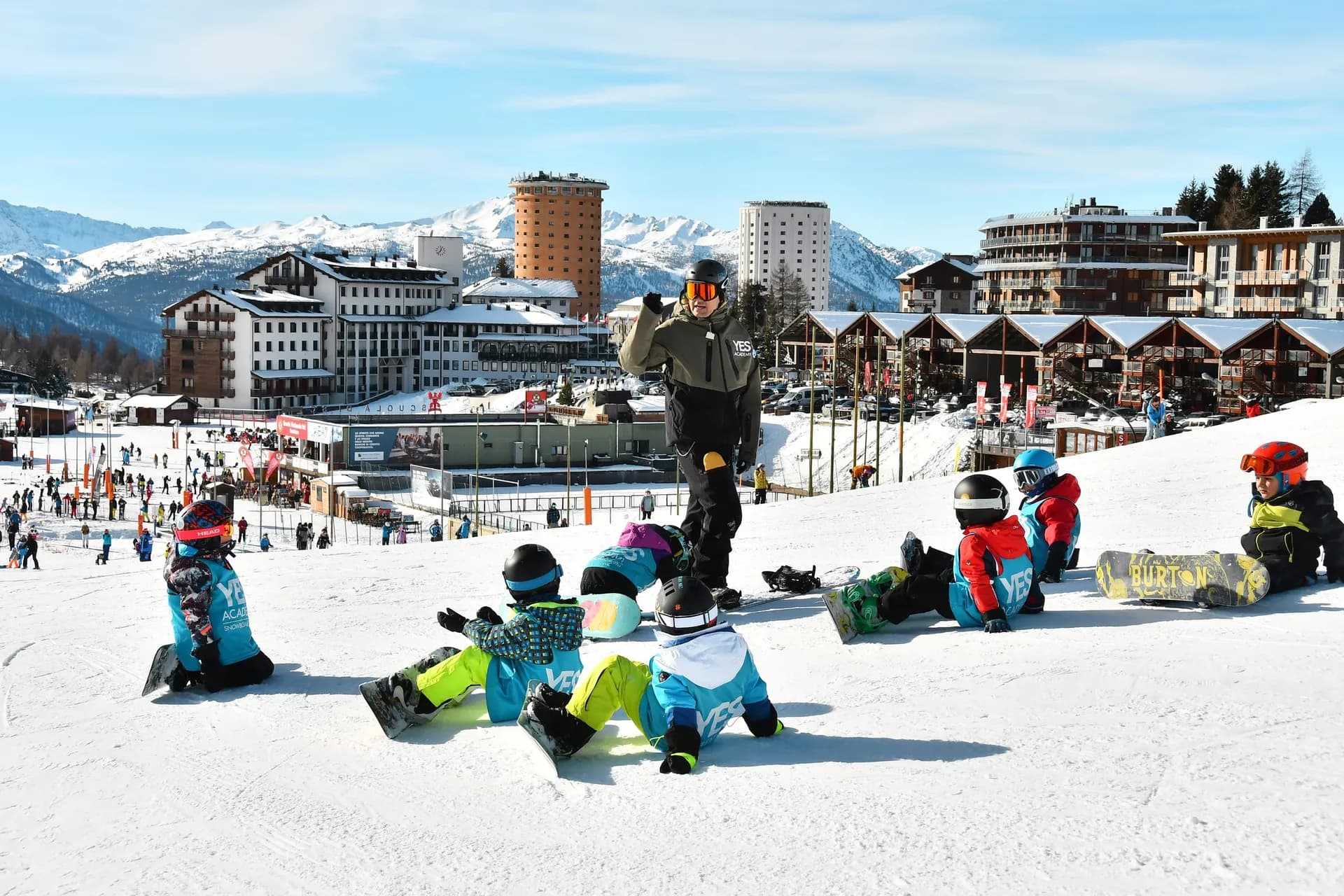 Maestro di snowboard parla a sette bambini seduti sulla neve con la tavola; dietro il paese di Sestriere e le Alpi