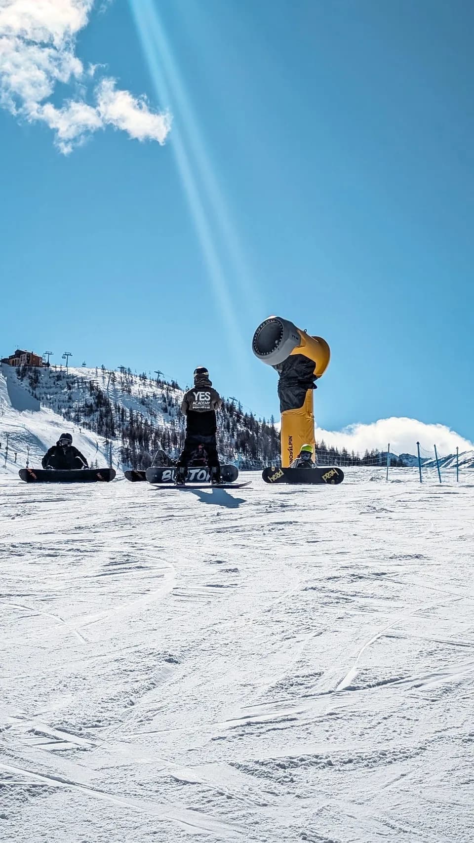 Maestro di snowboard di spalle davanti agli allievi seduti con la tavola, accanto a un cannone da neve giallo