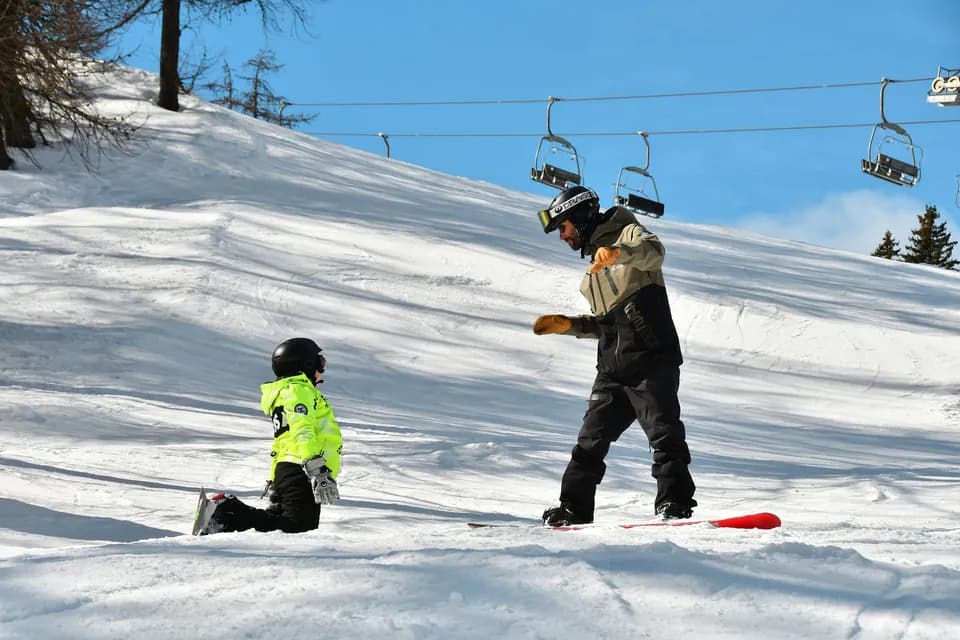 Maestro di snowboard mostra il movimento a un bambino seduto sulla neve con la tavola, seggiovia sullo sfondo